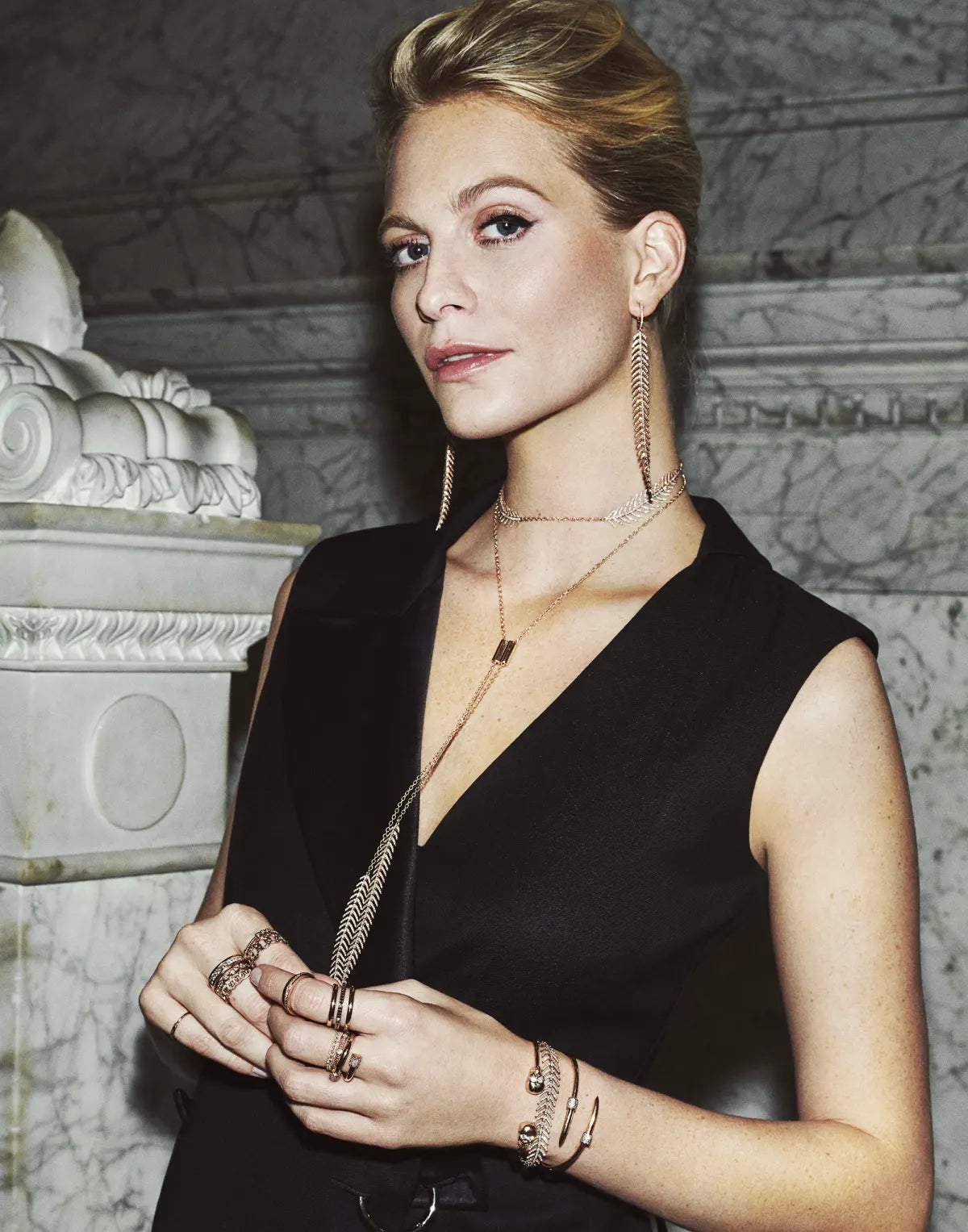A woman in a black sleeveless dress wearing layered Ilene Joy necklaces and rings beside a marble column